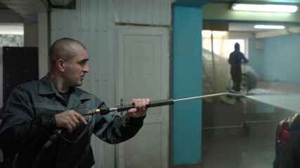 A Caucasian man works in a car wash service. A man in a worker's uniform holds a water gun and washes the car with a jet of water