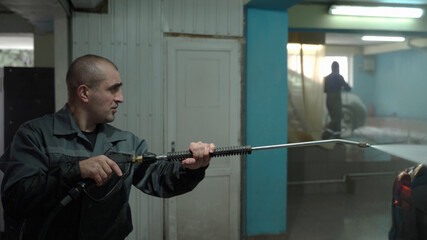 A Caucasian man works in a car wash service. A man in a worker's uniform holds a water gun and washes the car with a jet of water