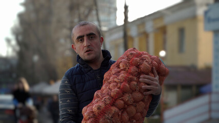 Close-up portrait of a Caucasian nationality male loader worker holding a large bag of onions in his hands