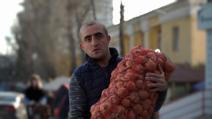 Close-up portrait of a Caucasian nationality male loader worker holding a large bag of onions in his hands