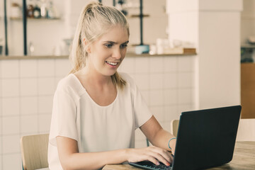 Happy beautiful blonde woman sitting at table in co-working space, using laptop, looking at display and smiling. Medium shot. Wireless communication or workplace concept