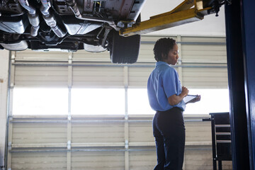 Female mechanic holding a digital tablets standing under a car chassis in auto repair shop