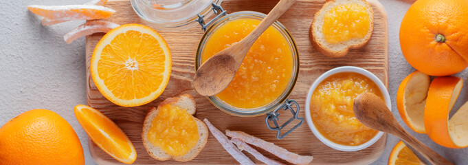 Orange jam in a jar, candied orange fruits, bread, fresh orange on a wooden board. Breakfast. Banner.