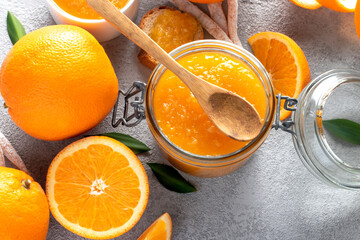 Orange jam in a glass jar with a spoon and fresh oranges, top view.