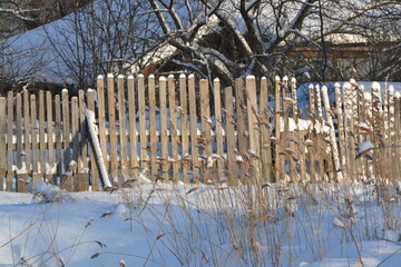Wooden fence with a locked door among snowdrifts and white snow near a house with a forest and...
