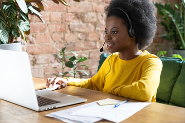 Focused Afro-American employee woman wearing headset, communicating via video call on laptop,...