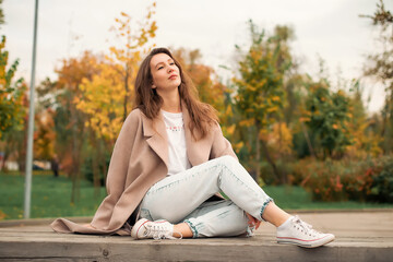 A girl with a Euro-Asian appearance in a pink coat and blue jeans with holes sits on a bench made of gray unpainted boards in a park