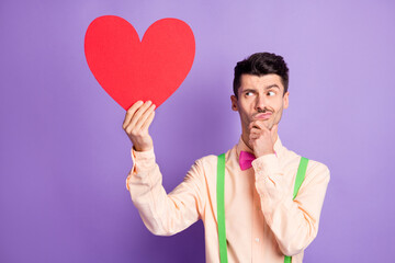 Photo of thoughtful unsure young man dressed yellow shirt holding large red paper heart arm chin isolated purple color background