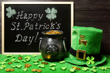 Happy St. Patricks Day chalk lettering on blackboard, leprechauns cast iron pot full of gold and hat, clover leaves and coins on green grass, dark wood. Saint Patricks Day banner, poster, invitation