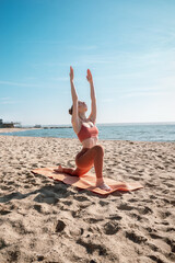 Young female in orange top and leggins practising Crescent Lunge Pose (Anjaneyasana) on a beach in the morning