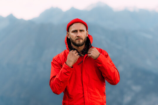 Portrait of a brutal bearded man in a red jacket and hat among the mountains. Male portrait on the background of a mountain landscape. A traveler on the background of mountains.