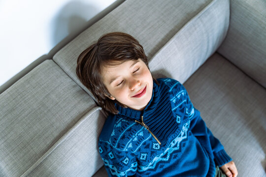 Portrait Of Young Boy Sitting On The Couch At Home, Threwing Back His Head And Closing Eyes With Pleasure. Child Relaxing And Dreaming On The Sofa After Long School Day.