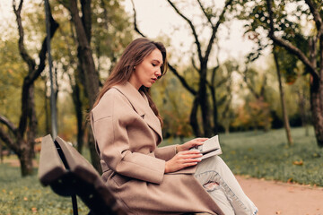 A Caucasian girl in a beige coat and blue jeans sits on a bench in a park in autumn and reads a book © Елизавета Шишова