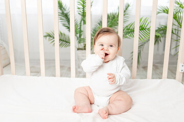 happy baby sitting in the crib in a white bodysuit and sucking his finger
