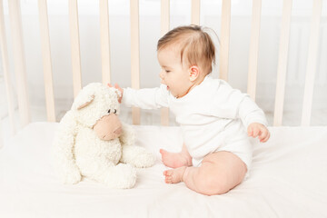 baby six months playing in a crib in a white bodysuit with a Teddy bear