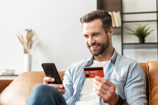 Bearded handsome man makes purchase online. A hipster wearing casual shirt holds a mobile phone and a credit card orders food. Smiling guy is using smartphone and debit bank card for paying in e-shop