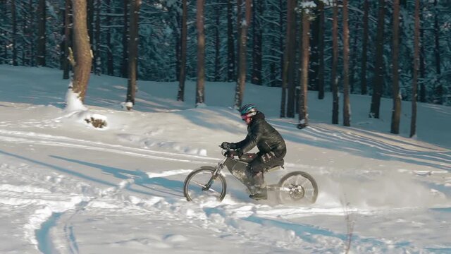 Cyclist in Black Clothes, Helmet and Snow Glasses Riding on Electric Bike in the Snow in Winter Forest. E-Bike Riding and Drifting at Cold Sunny Day - Slow Motion