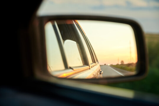 Close-up picture of car side mirror with beautiful road view with blue yellow sky at sunset. Auto traveling. Summer vacation road tripping.