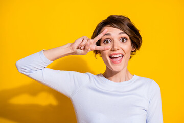 Photo portrait of cute girl showing v-sign near eye isolated on bright yellow colored background