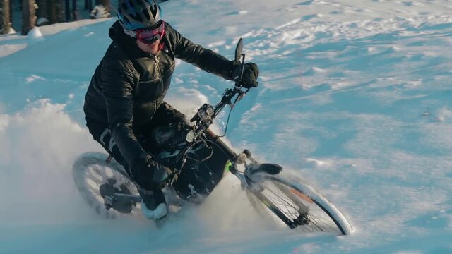 Cyclist in Black Clothes, Helmet and Snow Glasses Riding on Electric Bike in the Snow in Winter Forest. E-Bike Riding and Drifting at Cold Sunny Day - Slow Motion