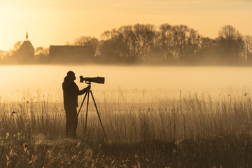 Photographer with a camera and telelens on a tripod photograping birds on a foggy morning. - obrazy, fototapety, plakaty