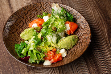 A salad with smoked salmon, mashed avocado and lettuce dressed with cream cheese. Close-up of the dish in a brown bowl isolated on wooden background. Horizontal orientation.
