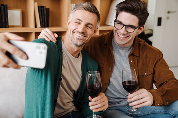 Happy gay couple drinking wine while taking selfie on cellphone