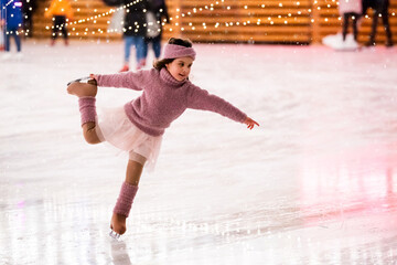Little girl figure skater in a pink sweater is skating on winter evening on an outdoor ice rink lit...