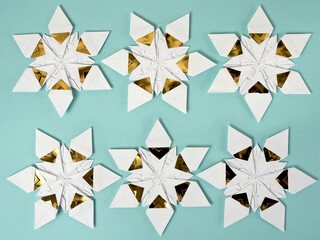 Six origami star snowflakes in two rows in white and gold foil paper, on a pale blue paper background