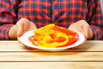 A girl holds a plate with chopped bell peppers on the background of a wooden table.