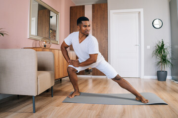 Front view of smiling strong African-American man doing side lunge exercise at home during working out standing on yoga mat at bright domestic room, looking away. Concept of sport training at home gym