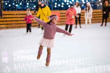 Little girl in pink sweater skates on winter snowy evening at an outdoor ice rink