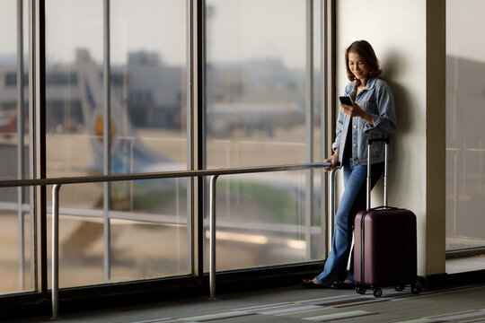 Beautiful Asian Traveler Standing Alone And Using Smartphone At The Airport While Waiting For A Flight