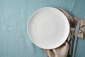 Clean empty white plate, fork and knife on pastel blue aquamarine linen tablecloth on table, copy space, mock up, top view. Concept for menu with utensil