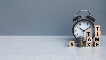 Tax time concept. An alarm clock with stack of money and wooden block with text on grey background with copy space.