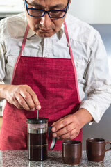 Vertical shot of man in red apron making fresh coffee in a French press next to two espresso cups. selective focus