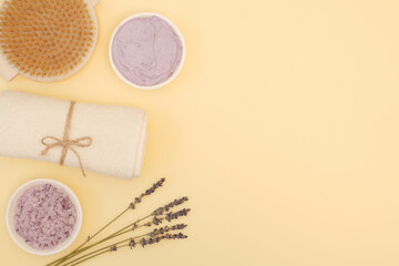 Fototapeta na wymiar Beautiful spa composition with a brush for dry anti-cellulite body massage, sea lavender bath salt, natural sea salt scrub, white towel, lavender sprigs on a yellow background with copy space. The