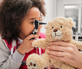 African American girl plays with her toy bear in a medical game, using an otoscope. Hearing clinic for children