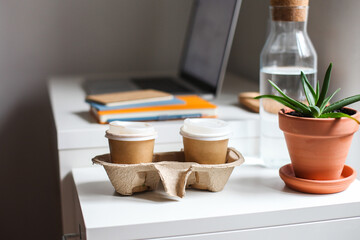 Two paper take away cups in a cardboard holder on a white table. Notebooks, laptop, workspace on the background