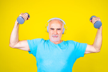 Elderly man training using dumbbells on yellow background smiling stretching using gym weight listening music