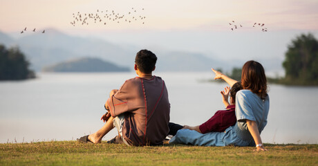 Mother and two sons sitting beside a big lake and see mountain view in the background, mom pointing...