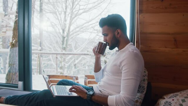 Freelancer, young indian man working on laptop and drinking hot chocolate while looking at snow covered trees. High quality 4k footage