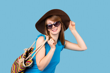 Happy blonde girl in hat and glasses with a beach bag posing on a blue background with side space. Vacation and travel concept.