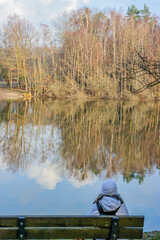Mature woman in winter clothes sitting on a wooden bench with her back to the camera, lake with mirror reflection in the water of the surrounding trees, sunny winter day in South Limburg, Netherlands