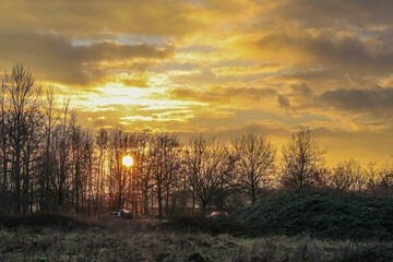 Dutch countryside with a beautiful sunset with the sun between the branches of the bare trees and the clouds on a cloudy afternoon, magical moment in Dal van de Roodebeek in South Limburg, Netherlands