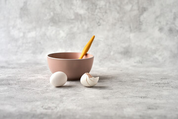 A ceramic bowl of delicate pink color with an egg and egg shell lying next to it on a gray background. Copy space.