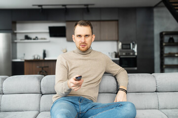 Fototapeta premium Handsome bearded young man holding tv remote control, switch channels, watching evening TV shows, spends leisure time on the couch at home in front of television set