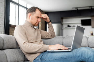 The guy working remotely has a headache, young man with laptop holding head, feels pain in...