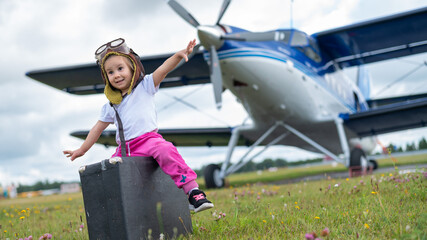A little girl in a pilot's costume carries a retro suitcase and walks along the airfield. A child...