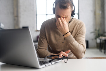 Portrait of a tired millennial young businessman in a headset sits at the desk with a laptop,...
